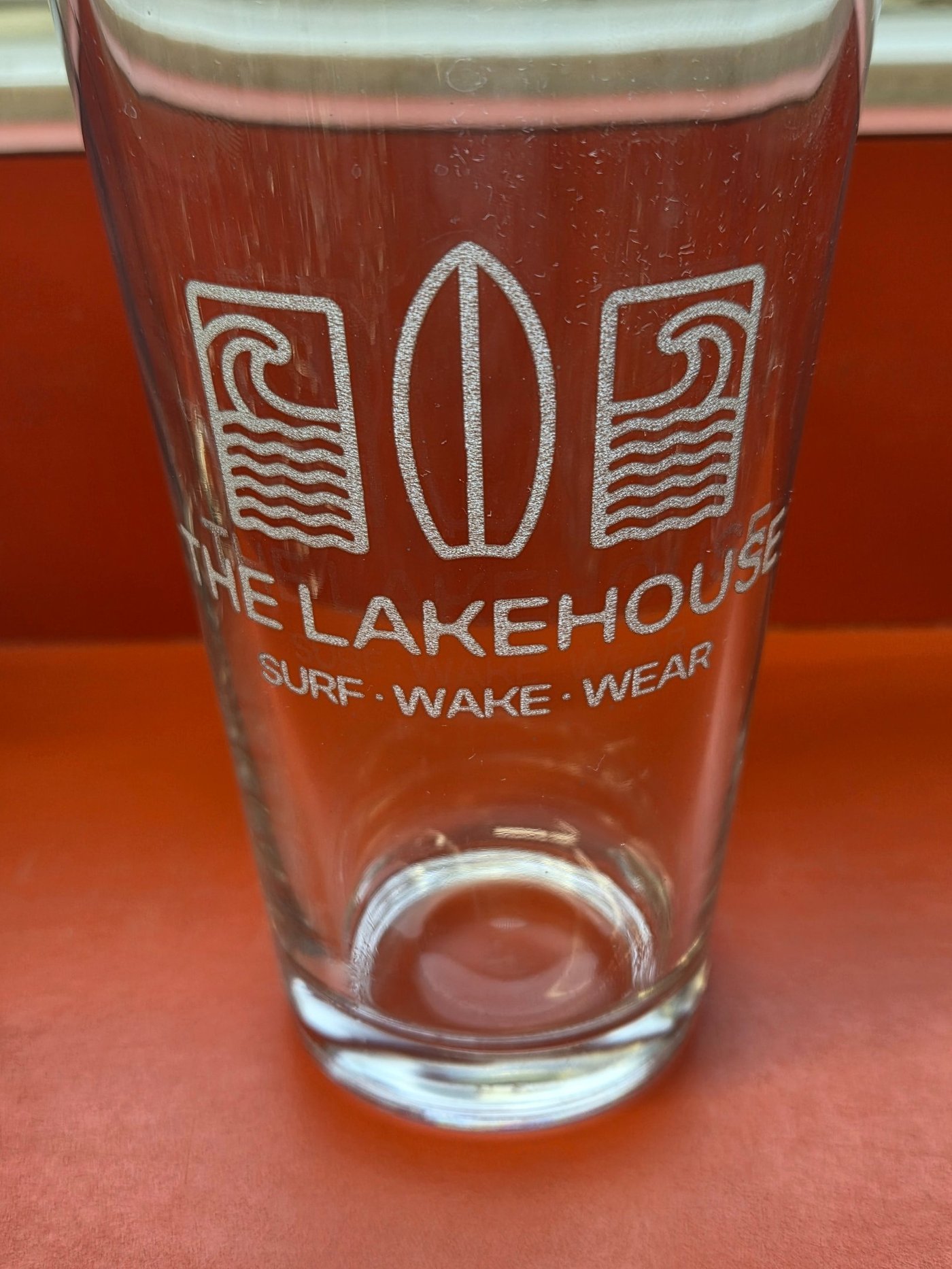 A clear pint glass frosted-etched with a surfboard and wave logo reading The Lakehouse, Surf Wake Wear.