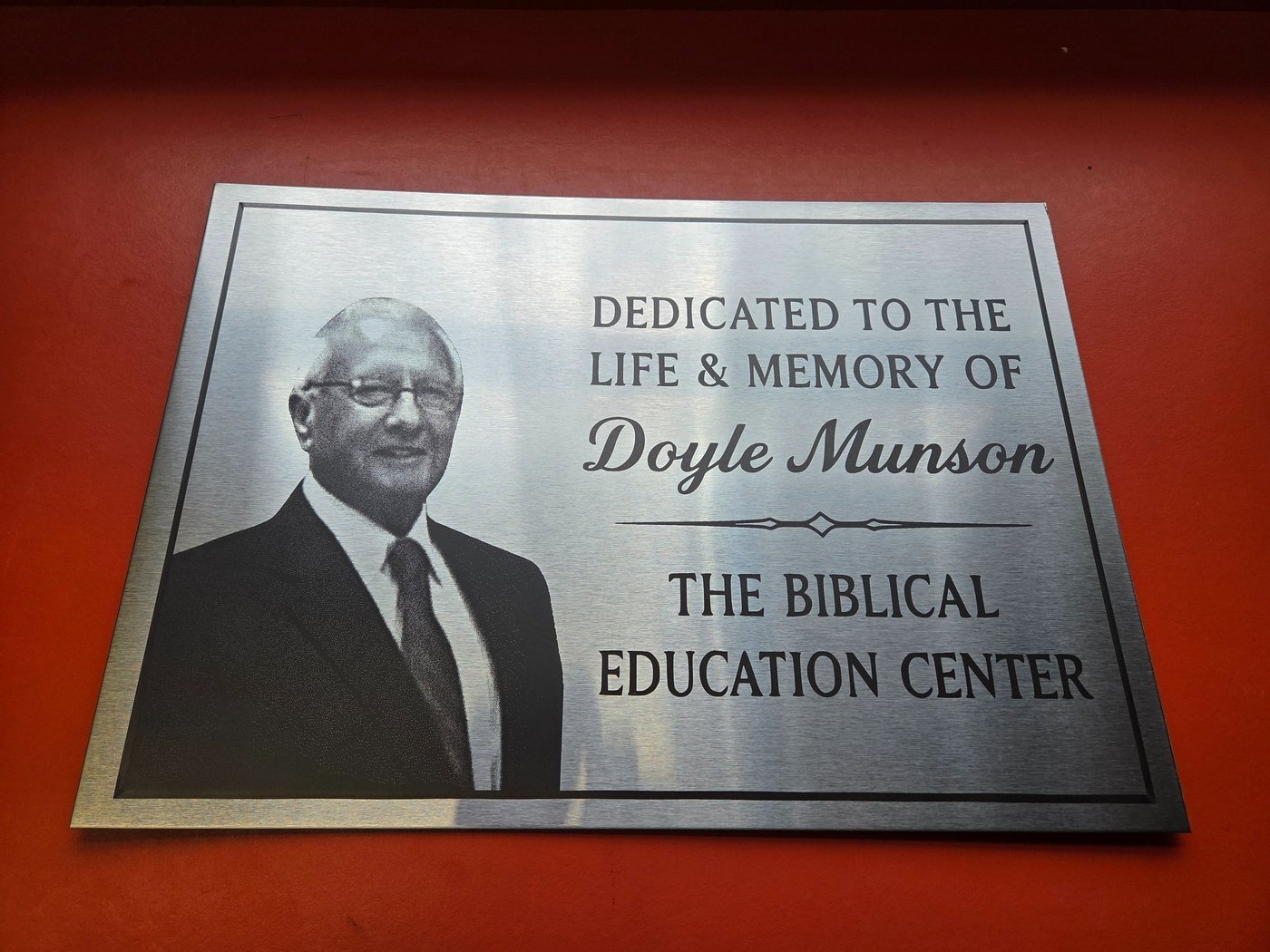 A brushed metal plaque engraved with a portrait photograph and the words Dedicated to the life and memory of Doyle Munson, The Biblical Education Center.