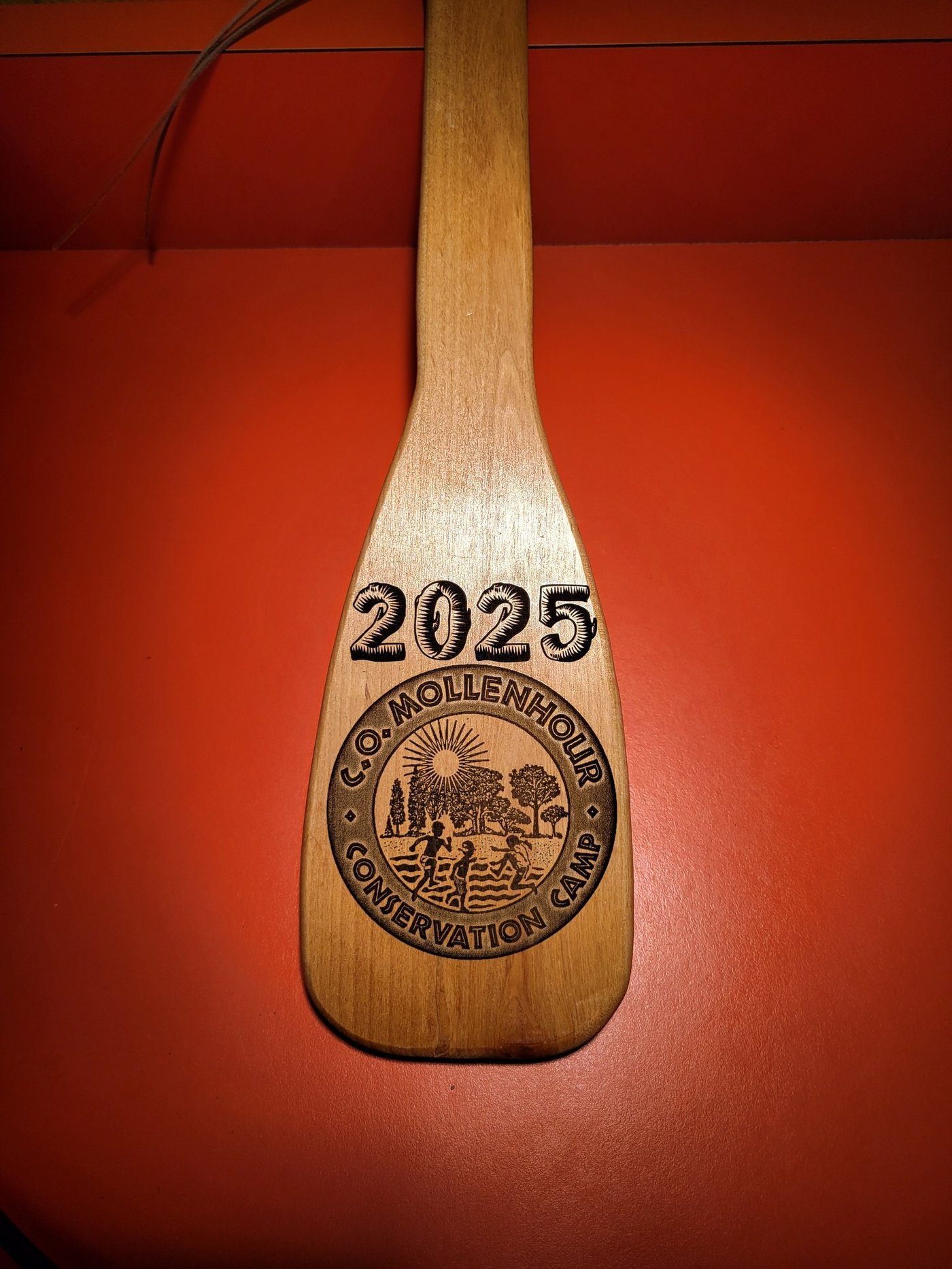 A wooden canoe paddle deeply engraved with a 2025 C.O. Mollenhour Conservation Camp emblem showing children, trees and a sunrise.