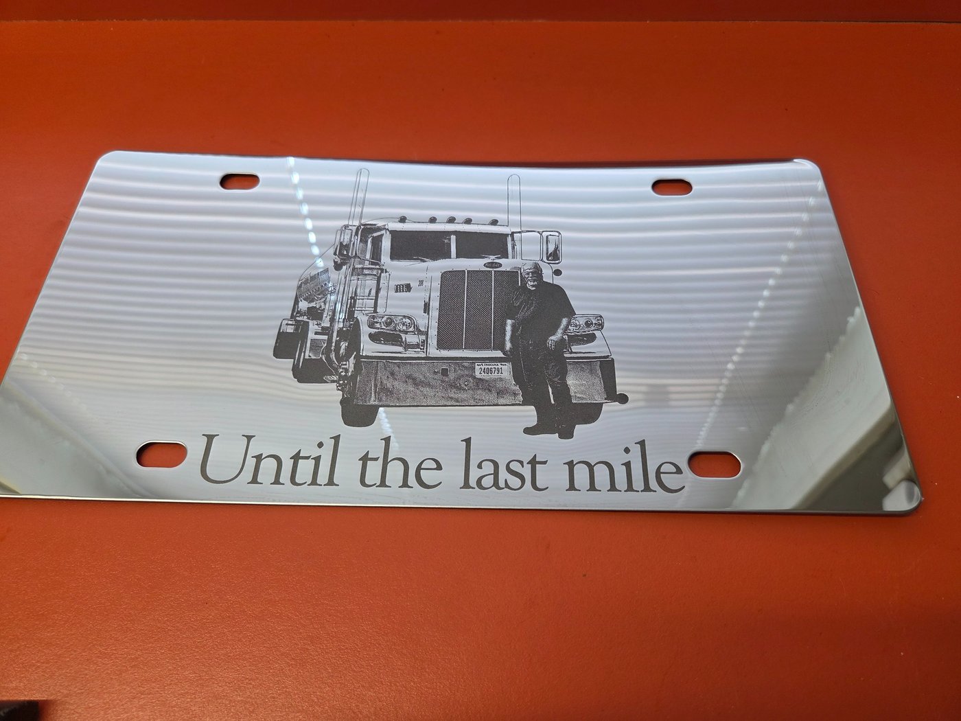 A white metal licence plate engraved with a semi truck and driver above the words Until the last mile.