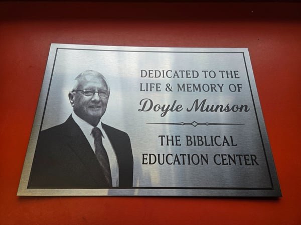 A brushed metal plaque engraved with a portrait photograph and the words Dedicated to the life and memory of Doyle Munson, The Biblical Education Center.