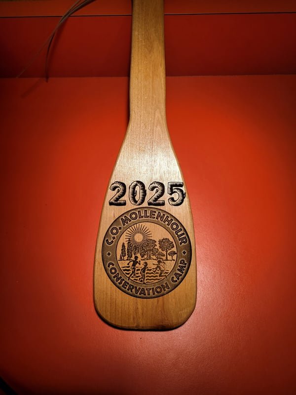 A wooden canoe paddle deeply engraved with a 2025 C.O. Mollenhour Conservation Camp emblem showing children, trees and a sunrise.