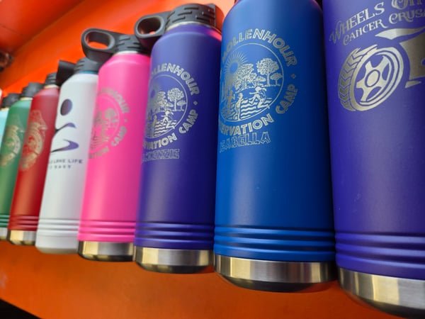 A row of insulated water bottles in white, pink, purple and blue, each engraved with a conservation camp or charity logo.