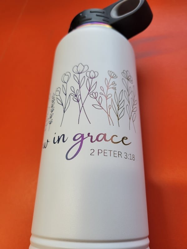 A white insulated bottle engraved with a wildflower illustration and the words Grow in grace, 2 Peter 3:18, the marks shifting from purple to bronze.