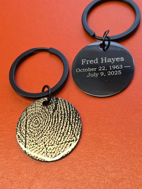 A black metal keyring tag engraved with Fred Hayes and his dates beside a gold disc bearing an engraved fingerprint.