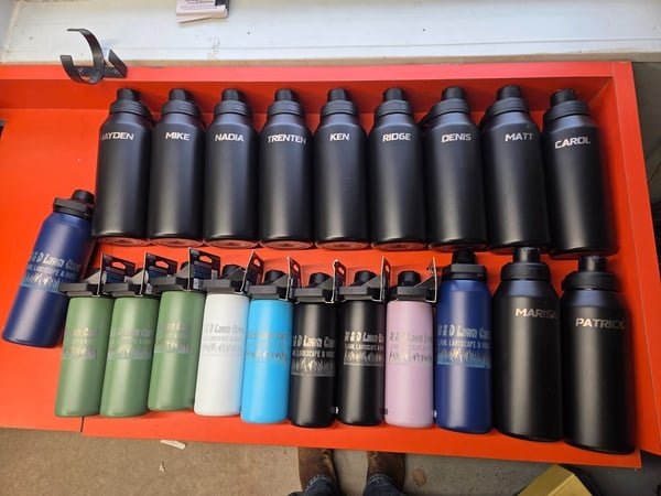 More than twenty insulated bottles laid out in rows, each engraved with a staff member's first name or a lawn-care company logo.