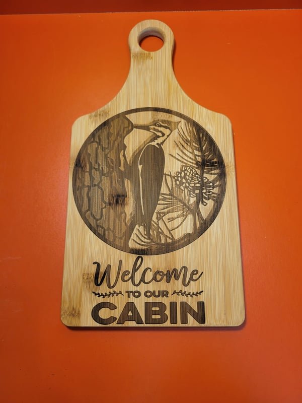 A bamboo cutting board engraved with a woodpecker on a tree and the words Welcome to our Cabin.