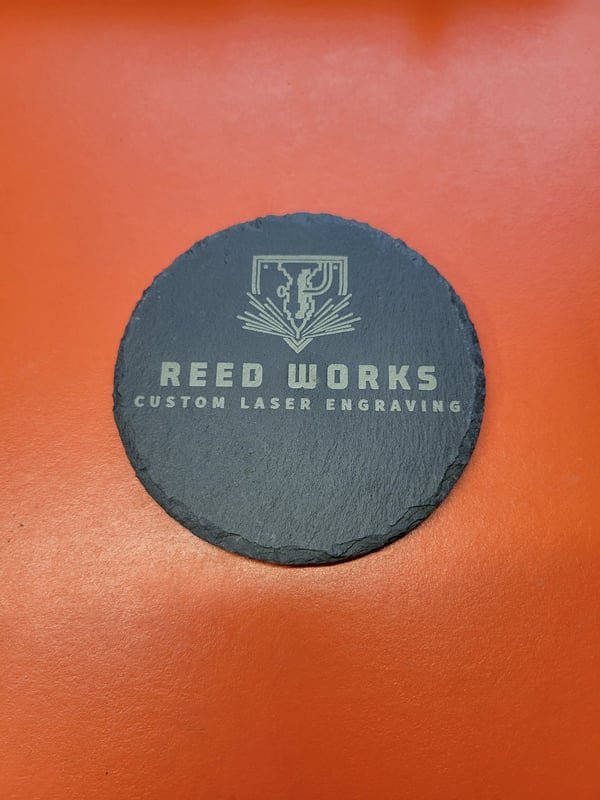 A round slate coaster engraved with the Reed Works Custom Laser Engraving logo.