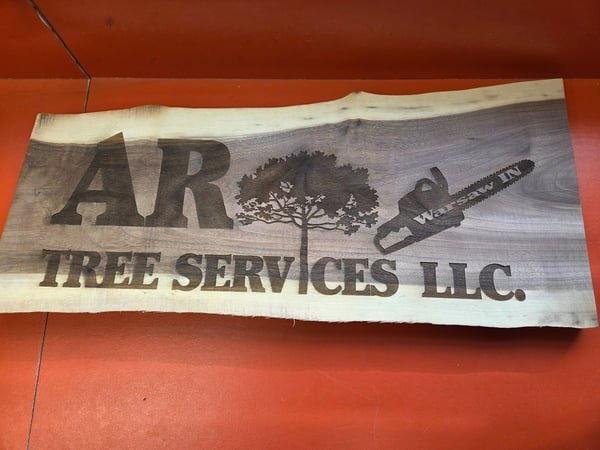 A live-edge wood sign engraved with the AR Tree Services LLC logo, a tree and a chainsaw.