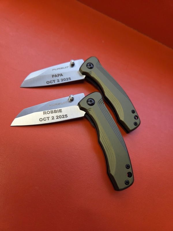 Two folding knives with green handles, their blades fiber-engraved PAPA and ROBBIE with the date Oct 2 2025.