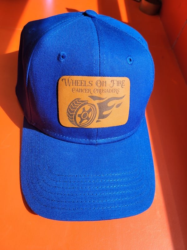 A blue cap with a leather patch engraved with the Wheels On Fire Cancer Crusaders flaming-wheel logo.