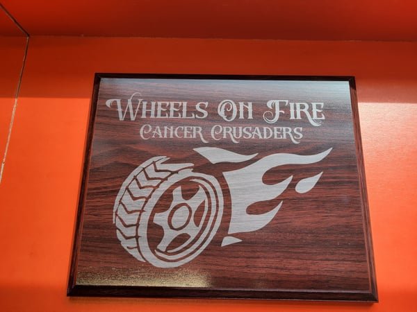 A dark wood plaque engraved with the Wheels On Fire Cancer Crusaders flaming-wheel logo.
