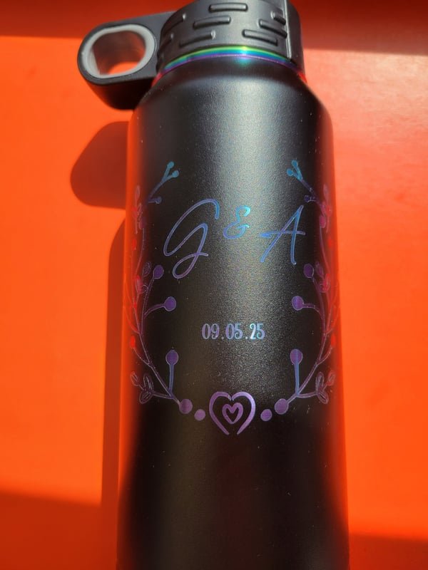 A dark tumbler engraved with the monogram G and A and the date 09.05.25, the marks shifting purple and gold.