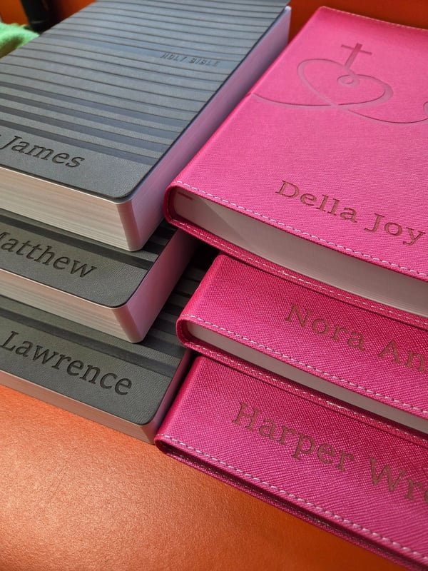 A stack of leather-bound notebooks in grey and pink, each engraved with a different person's name.