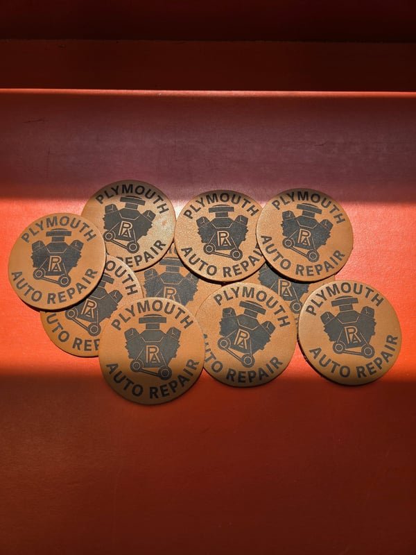 A pile of round leather patches engraved with the Plymouth Auto Repair logo.