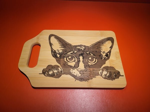 A bamboo cutting board engraved with a wide-eyed cat peering over the edge.