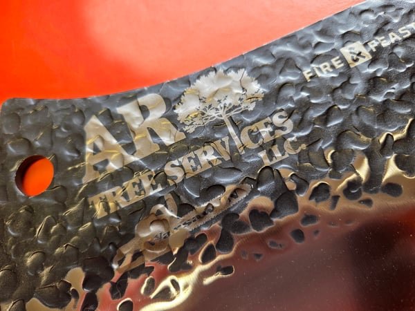 A hammered steel cleaver blade fiber-engraved with the AR Tree Services logo.