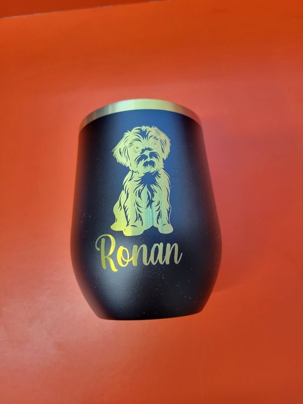 A black stemless wine tumbler engraved with a portrait of a shaggy dog and the name Ronan.