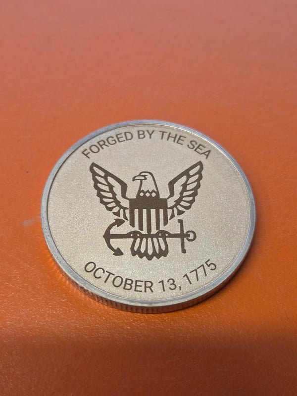 A metal challenge coin fiber-engraved with an eagle and the words Forged By The Sea, October 13, 1775.