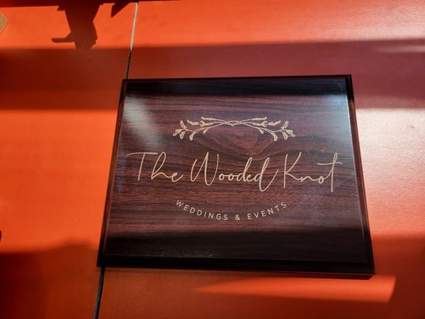 A dark plaque engraved with The Wooded Knot, Weddings and Events, above a floral flourish.