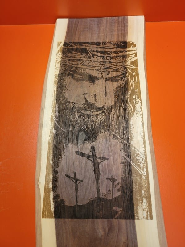 A live-edge wood board engraved with a detailed crucifixion scene and the face of Christ.