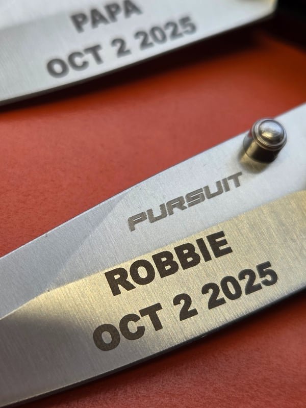 A stainless knife blade fiber-engraved Pursuit, Robbie, Oct 2 2025.