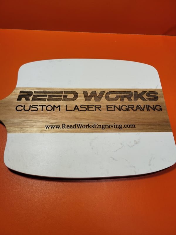 A marble and wood serving board engraved with the Reed Works logo and website.