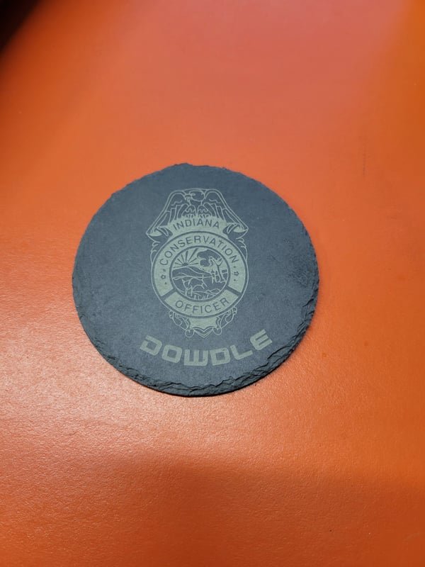 A round slate coaster engraved with the Indiana Conservation Officer badge and the name Dowdle.