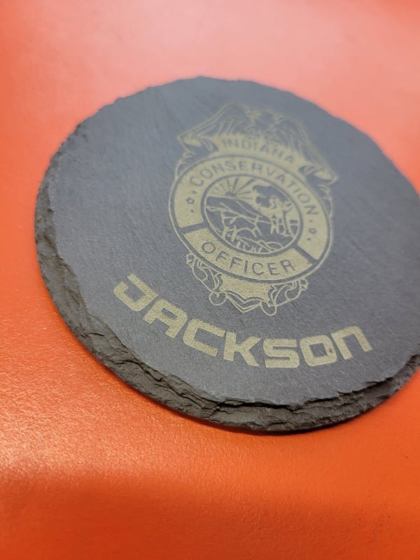 A round slate coaster engraved with the Indiana Conservation Officer badge and the name Jackson.