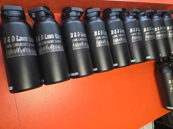 A row of black insulated bottles engraved with the H&D Lawn Care, Landscape and More logo.