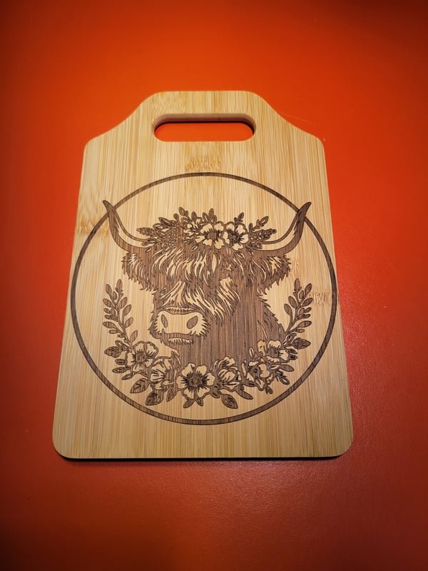 A bamboo cutting board engraved with a highland cow framed by a floral wreath.