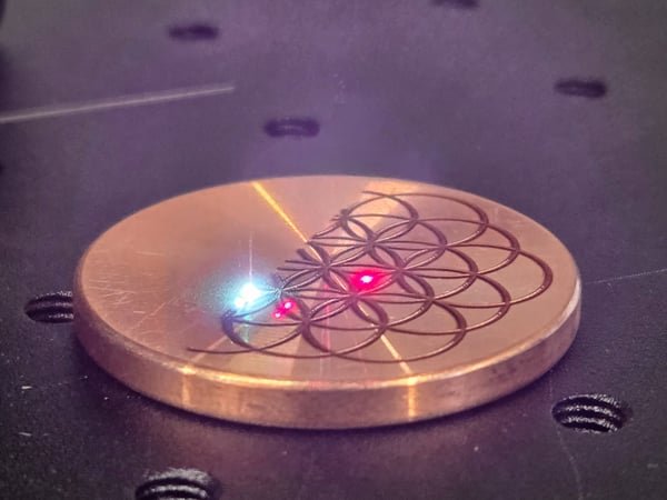 A copper disc on the laser bed mid-job, the beam tracing a geometric pattern into its face.