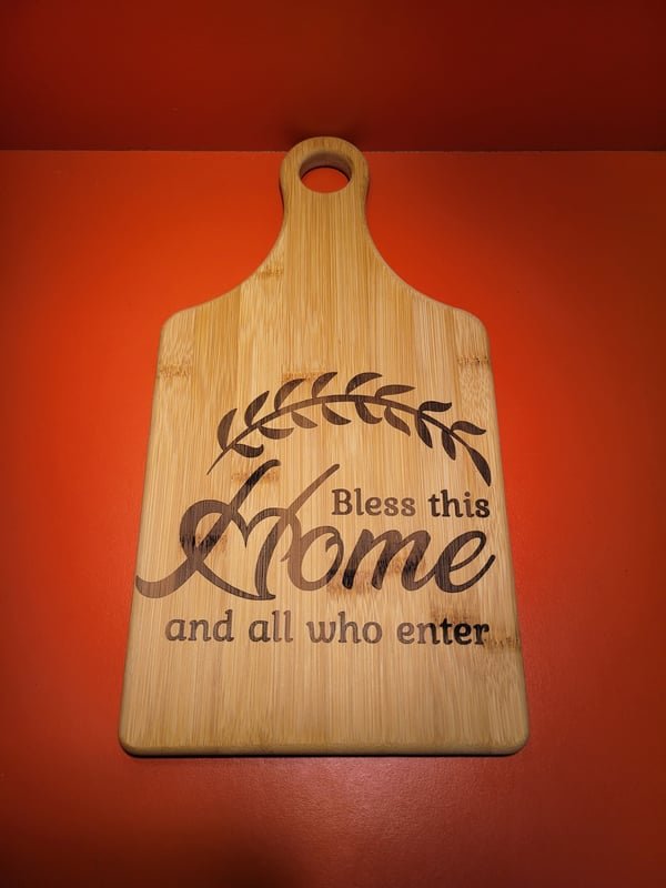 A bamboo paddle board engraved with a laurel wreath and the words Bless this Home and all who enter.