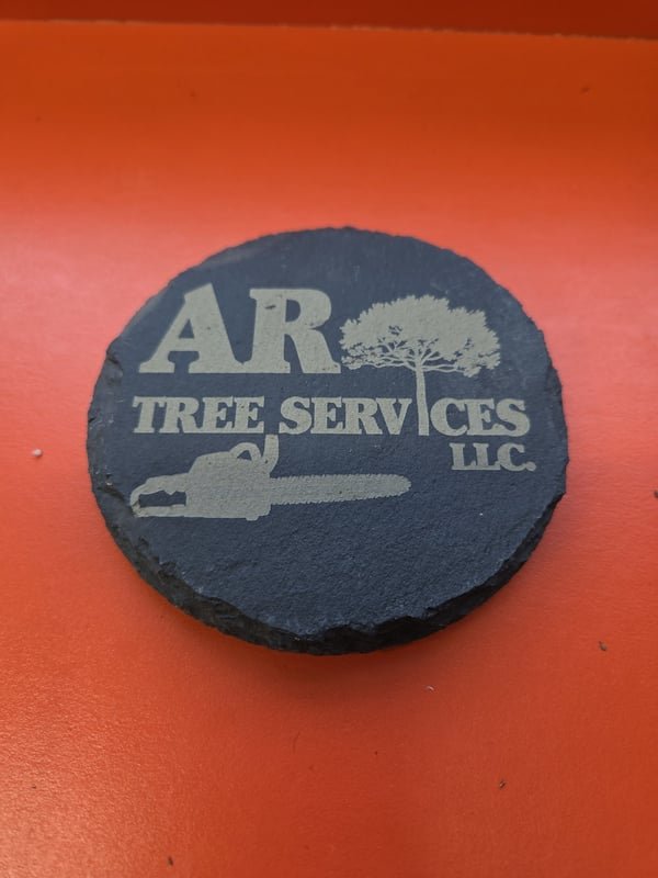 A round slate coaster engraved with the AR Tree Services logo and chainsaw.