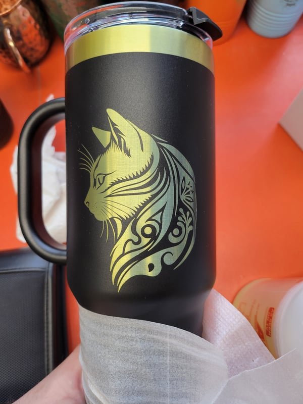 A black tumbler engraved with a stylised tribal cat in gold-toned marks.