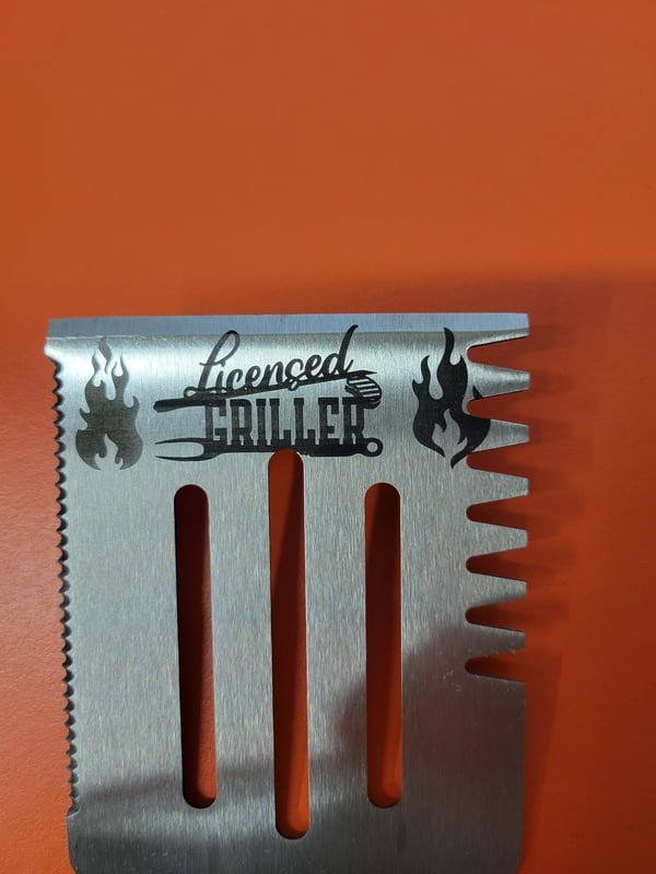 A stainless grill scraper fiber-engraved with flames and the words Licenced Griller.