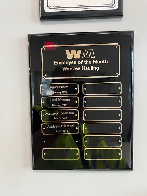 A wall-mounted recognition board fitted with engraved brass plates naming each Employee of the Month for Warsaw Hauling.