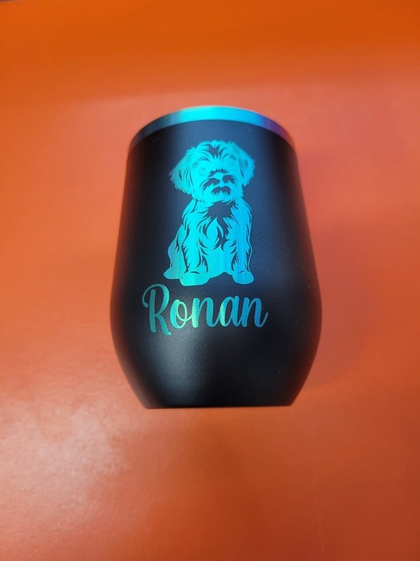A black stemless wine tumbler engraved with a shaggy dog portrait and the name Ronan in teal.
