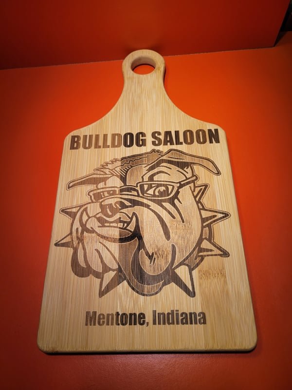 A bamboo paddle board engraved with a bulldog mascot reading Bulldog Saloon, Mentone, Indiana.