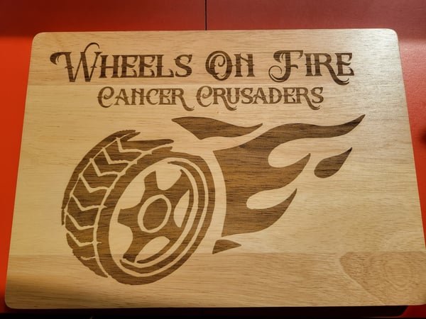 A wood sign engraved with the Wheels On Fire Cancer Crusaders flaming-wheel logo.