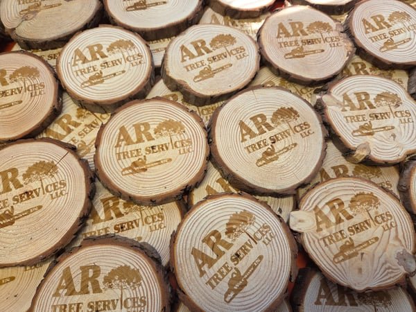 A large batch of wood slice coasters, each engraved with the AR Tree Services logo.