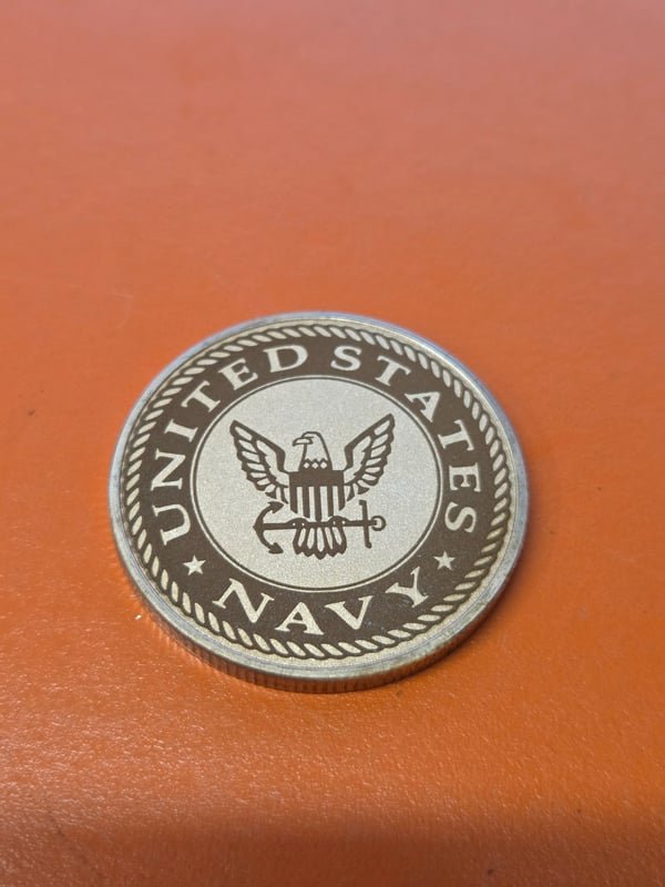 A metal challenge coin fiber-engraved with the United States Navy eagle and anchor crest.