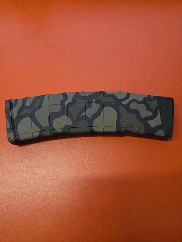 A rifle magazine in grey camouflage, fiber-engraved along its curved body.