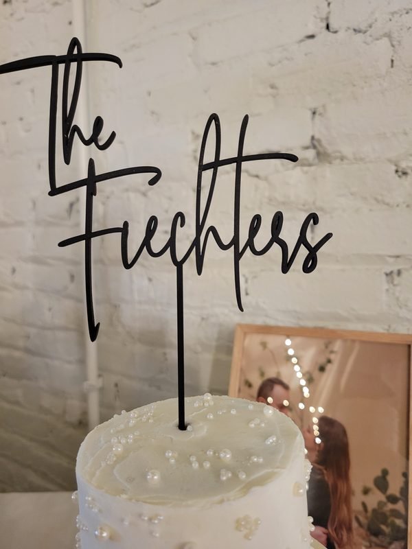 A black acrylic cake topper cut in flowing script reading The Fiechters, standing in a pearl-dressed wedding cake.