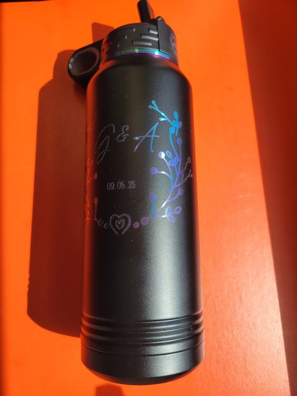 A black insulated bottle engraved with the monogram G and A, the date 09.05.25 and a floral spray in colour-shifted marks.
