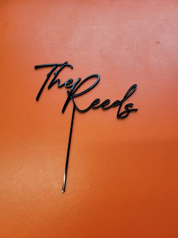A black acrylic cake topper cut and engraved in flowing script reading The Reeds.