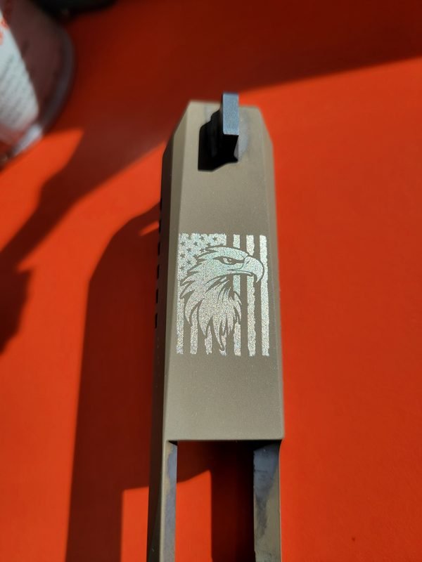A close-up of a tan pistol slide, the eagle and flag engraving catching the light.