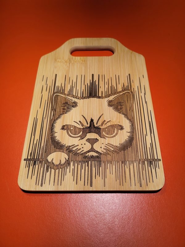 A bamboo cutting board engraved with a grumpy-faced cat framed by fine vertical lines.