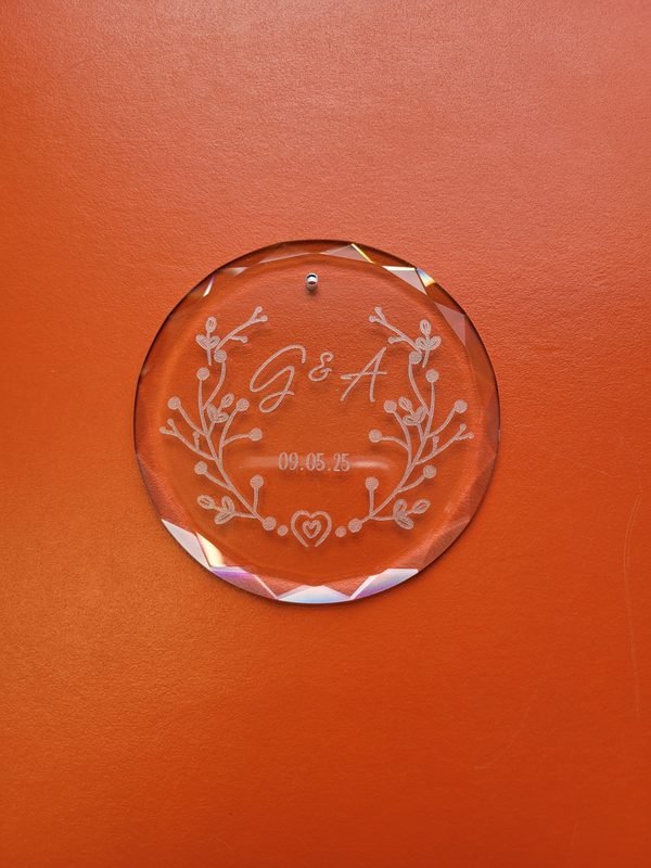 A round faceted glass ornament engraved with the monogram G and A, the date 09.05.25 and a floral wreath.