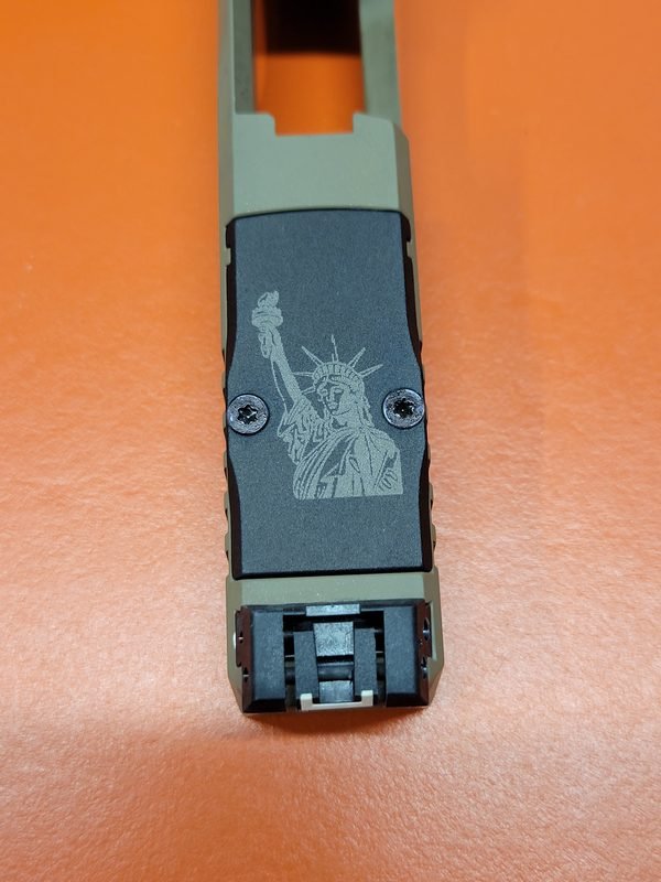 An olive drab pistol slide fiber-engraved with the Statue of Liberty holding her torch.